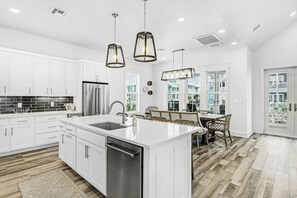 A modern kitchen with white cabinetry, a stainless steel refrigerator, and a large island with a sink. - Sunset Avenue Stunner | Steps to Beach (Port Aransas)