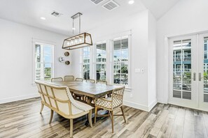 A dining area with a large wooden table and wicker chairs, large windows, and a view of a residential area. - Sunset Avenue Stunner | Steps to Beach (Port Aransas)