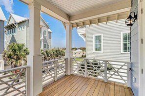 Une terrasse en bois couverte avec des balustrades blanches, un bâtiment de couleur claire doté de volets et un ciel dégagé.