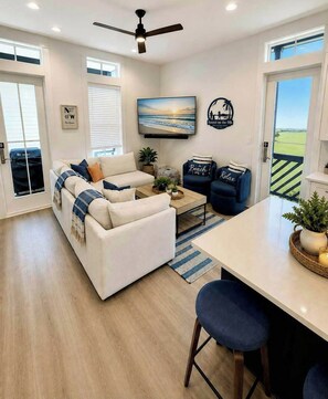 A modern living room with a sofa, coffee table, and two armchairs. There is a painting on the wall, a ceiling fan, and a view of the outdoors. - Sunset On The 9th | Golf Views | Steps to Beach (Port Aransas)