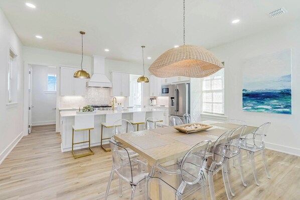 A modern kitchen with white cabinetry, a central island, and a dining area with clear chairs. - Sunsets And Smiles| Resort and Beach Access (Port Aransas)