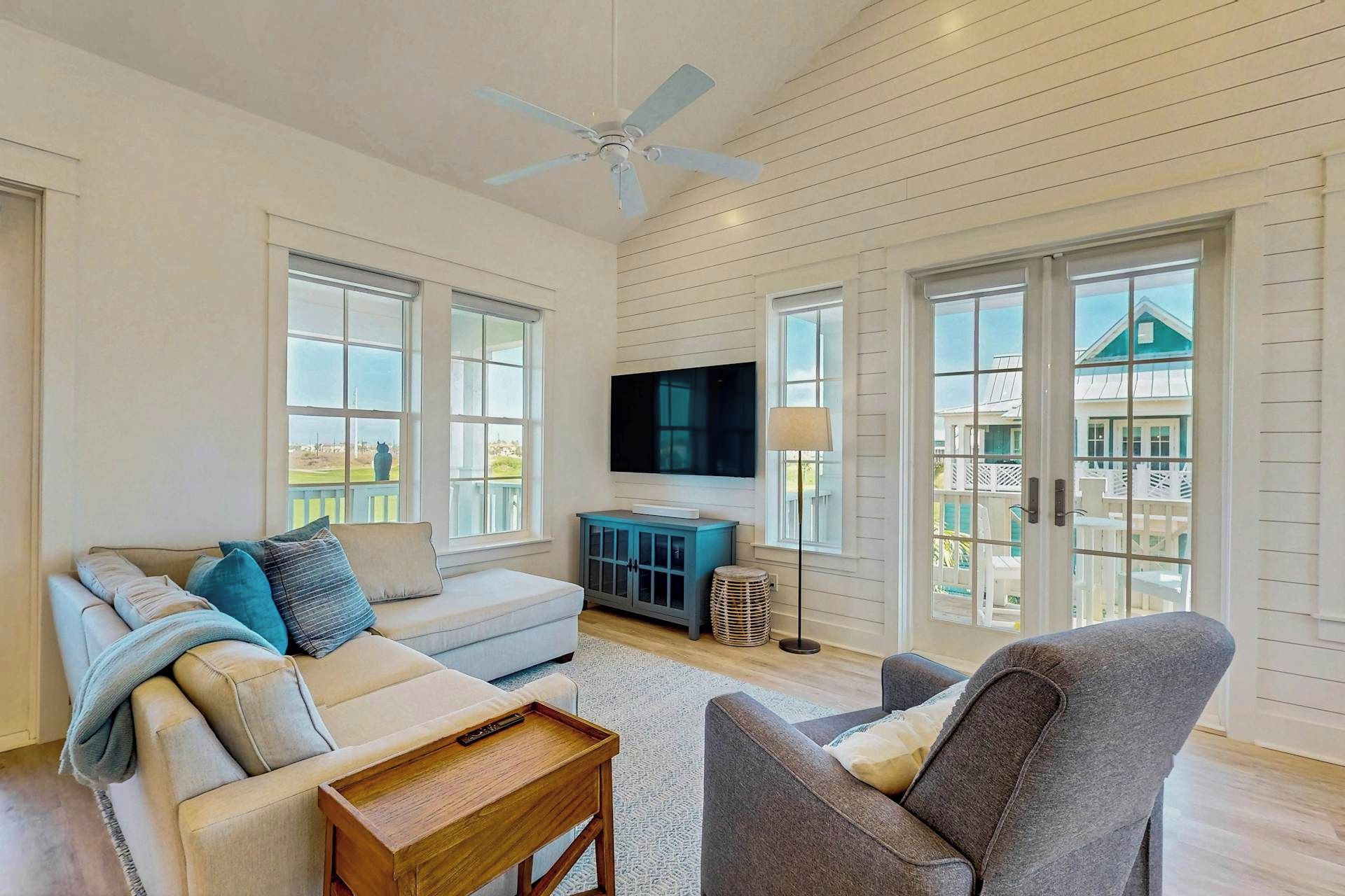 A spacious living room with a ceiling fan, large windows, a flat-screen TV, and a view of a house outside.