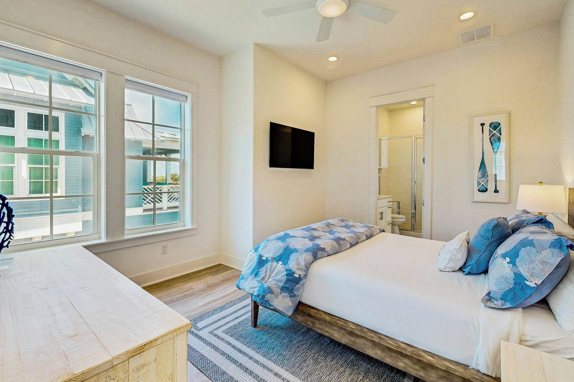 A bedroom with a bed, a desk, a chair, a ceiling fan, a TV, a mirror, and a window with a view of a house and a fence.
