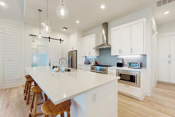 A modern kitchen with white cabinetry, a large island, stainless steel appliances, and pendant lighting. - The Beach Ball | Golf Views | Steps to Beach (Port Aransas)