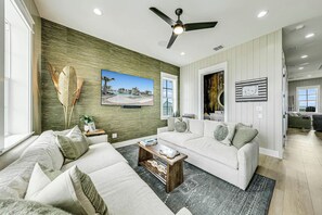 A living room with a large flat-screen TV, a ceiling fan, and a view of the ocean through the window. - The Palms | Private Elevator | Steps to Beach (Port Aransas)