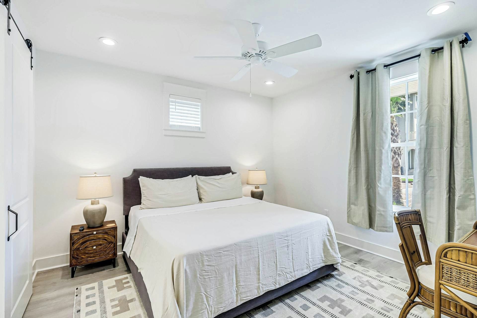 A bedroom with a bed, two bedside tables, a ceiling fan, a window with curtains, and a view of the outdoors.