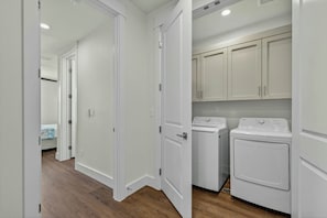 A laundry room with two white washing machines and a dryer, white cabinets above, and a wooden floor. - Palmilla Blue Ocean Blessing | Steps to Beach (Port Aransas)