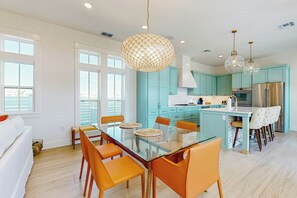 A modern kitchen with teal cabinets, a central island, and a dining area with orange chairs and a glass table. - Sip Sip Hooray | Steps to Beach | Resort Amenities (Port Aransas)