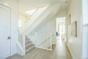 A staircase with white railings leading to an upper floor, a door on the left, and a hallway with a patterned floor and a door at the end. - Sip Sip Hooray | Steps to Beach | Resort Amenities (Port Aransas)