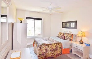 Une chambre d’hôtel comprenant un lit, un bureau, une chaise, une fenêtre, un ventilateur de plafond et une table de chevet avec une lampe.