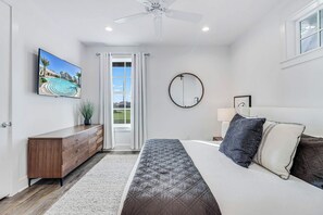 Ein Schlafzimmer mit einem Bett, einer Kommode, einem Deckenventilator, einem Fenster mit Vorhängen, einem wandmontierten Spiegel und einem Gemälde von einem Schwimmbad.