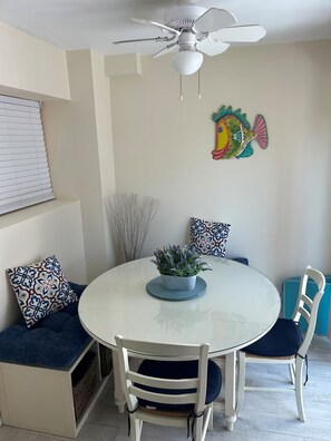 A dining area with a round table, white chairs, a ceiling fan, and decorative pillows.