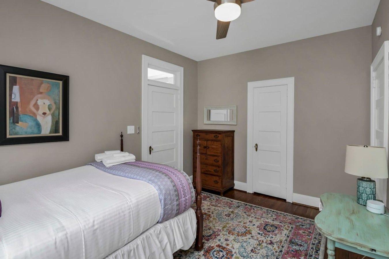 A bedroom with a bed, two doors, a dresser, a ceiling fan, and a painting on the wall.