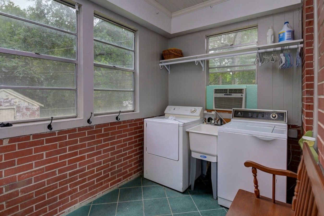 A laundry room with two washing machines, a dryer, and a utility sink. There are windows on the left and a shelf with cleaning supplies on the right.
