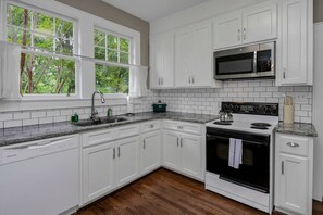 Una cocina con gabinetes blancos, un microondas de acero inoxidable, una estufa y una ventana con vistas a árboles.