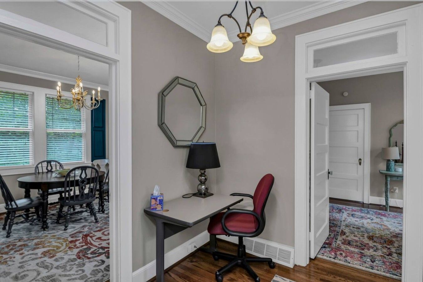 A room with a dining table, chairs, a desk with a chair, a lamp, a mirror, and a patterned rug.
