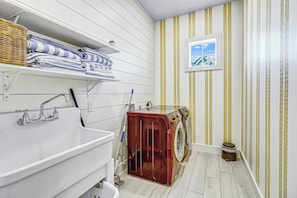 A bathroom with a white sink, a wooden washing machine, and a window with a view of a house. - Sunny Daze | Gulf Views | Steps to Beach | Firepit (Port Aransas)