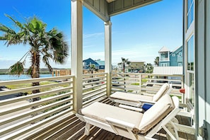 A covered outdoor seating area with lounge chairs and a view of a coastal town. - Sunny Daze | Gulf Views | Steps to Beach | Firepit (Port Aransas)