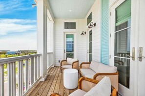A covered porch with wicker furniture, a round white table, and a view of a distant house. - Sunset Hacienda | Water Views | Steps to Beach (Port Aransas)