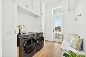 A laundry room with two front-loading washing machines, white cabinetry, and a view of a blue house through a glass door. - Sunset Hacienda | Water Views | Steps to Beach (Port Aransas)