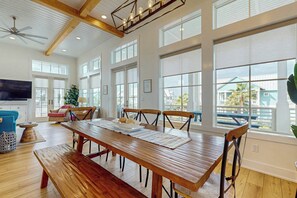 A spacious dining area with a long wooden table, multiple chairs, and large windows offering views of the outside. - Sunset Palms | Steps to Beach | Putting Green (Port Aransas)