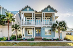 A two-story house with a front porch, a balcony on the upper floor, and palm trees in the front yard. - Sunset Palms | Steps to Beach | Putting Green (Port Aransas)
