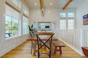 A spacious dining area with a long wooden table, chairs, and a white railing along one side. - Sunset Palms | Steps to Beach | Putting Green (Port Aransas)