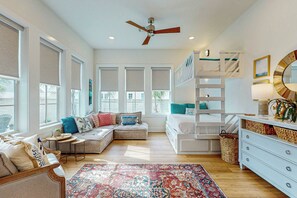 A spacious room with a large window, a ceiling fan, a bunk bed, a white dresser, and a patterned rug. - Sunset Palms | Steps to Beach | Putting Green (Port Aransas)