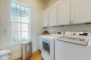 A laundry room with a dryer, washing machine, and a trash can. - Sunset Palms | Steps to Beach | Putting Green (Port Aransas)