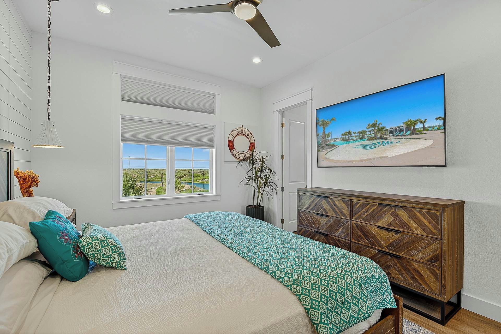 Een slaapkamer met een bed, een plafondventilator, een raam met uitzicht, een dressoir en een schilderij van een strand.