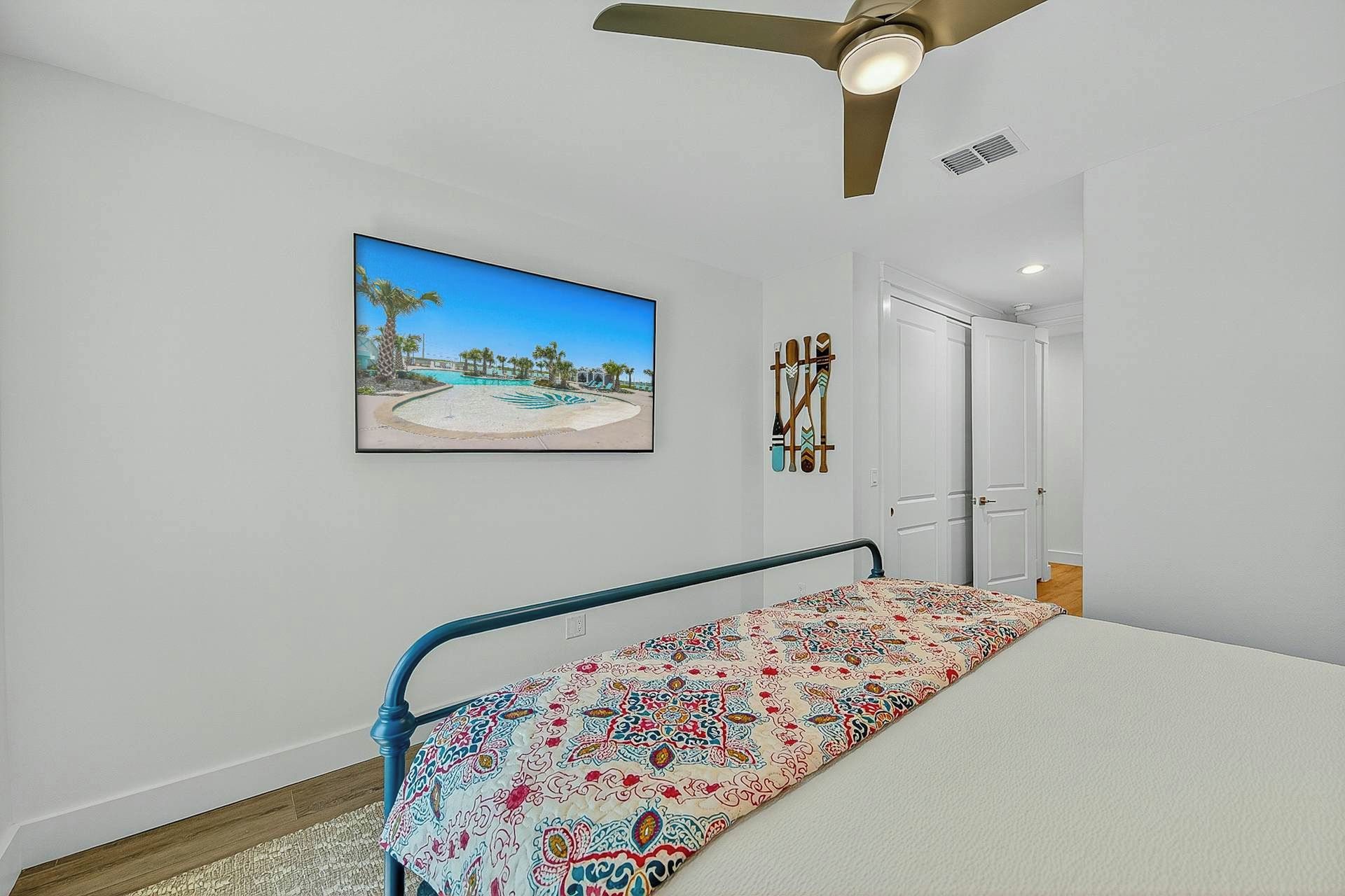 Une chambre à coucher avec un lit, un ventilateur de plafond, une porte et un tableau représentant une scène de plage, accroché au mur.