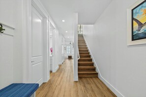 Ein Flur mit einer Holztreppe, die zu einer oberen Etage führt, weißen Wänden und einem Gemälde an der rechten Wand.