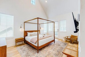 Een kamer met een slaapkamer met hemelbed, een houten frame, nachtkastje, lamp en stoel, grote ramen en een schuine kap.