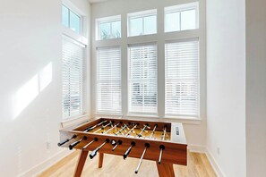 A room with a foosball table, large windows with blinds, and a wooden floor. - Sunshine Overtime | Water Views | Private Pool (Port Aransas)