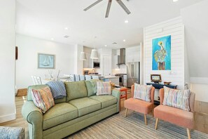 A modern living room with a green sofa, striped armchairs, and a kitchen in the background. - The Coral Monkey | Resort Amenities (Port Aransas)