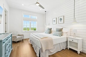 A bedroom with a large bed, a nightstand, a dresser, a chair, a window with a view, and a ceiling fan. - The Coral Monkey | Resort Amenities (Port Aransas)