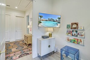 A hallway with a television displaying a pool, a patterned rug, a white console, and children's books on the wall. - The Palm | Steps to Beach | Resort Amenities (Port Aransas)