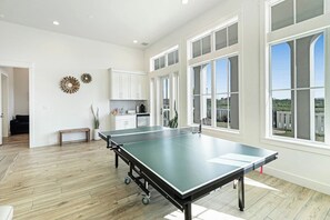 A spacious room with a ping pong table, large windows, and a kitchen area. - Pelican's Perch | Private Elevator | Putting Green (Port Aransas)
