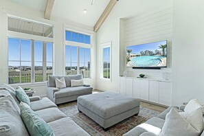 A spacious living room with a large flat-screen TV mounted on the wall, a grey sectional sofa, and a patterned rug. - Pelican's Perch | Private Elevator | Putting Green (Port Aransas)