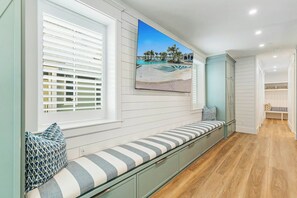 A room with a built-in bench, a window with blinds, a painting of a beach, and a bench with cushions. - Palmilla Pearl-Tallest In Palmilla | Gulf Front (Port Aransas)
