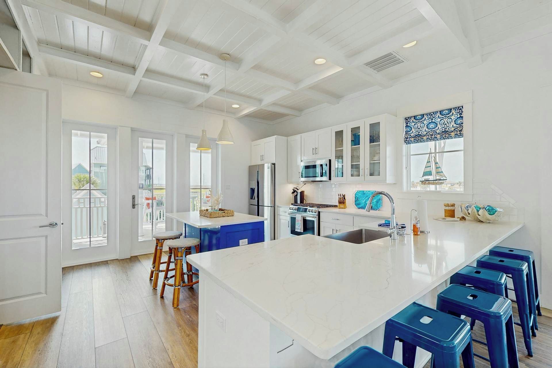 A modern kitchen with white cabinetry, a large island with blue bar stools, and a view of the outdoors through large windows.