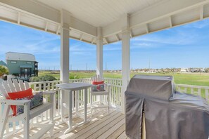 Ein überdachter Außenbereich mit weißer Möbel, einem überdachten Grill und Blick auf eine Wohngegend und den Strand.