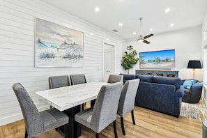 Ein modernes Wohnzimmer mit Essbereich, ausgestattet mit einer weißen Wand mit horizontalen Linien, einem Deckenventilator, einem Flachbildfernseher und einer großen Pflanze.