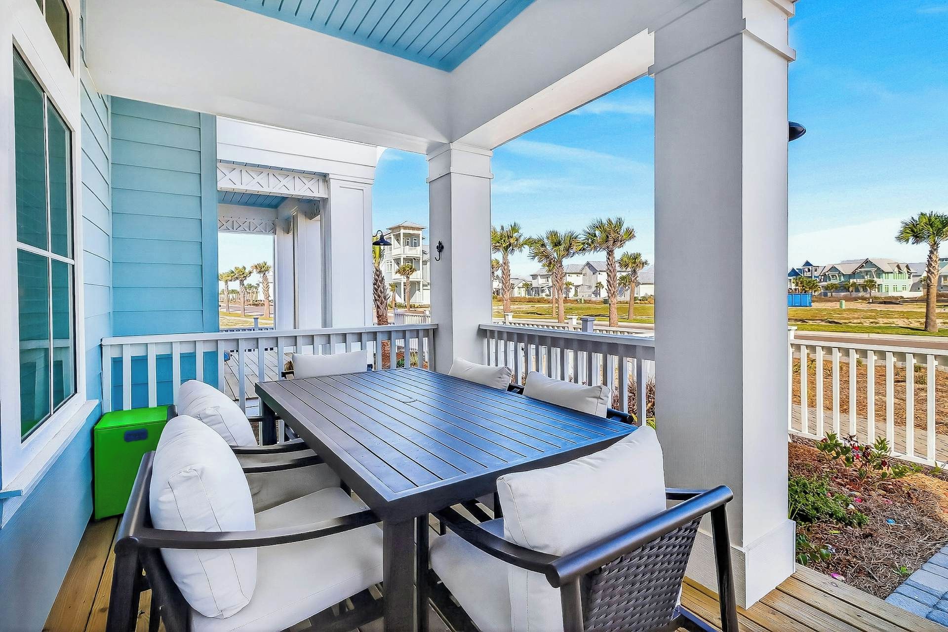 A covered outdoor seating area with a table and chairs, overlooking a residential neighborhood with palm trees and houses.