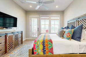 A bedroom with a bed, a TV, a ceiling fan, and a view of the outside. - Indigo Mist | Steps to Beach | Resort Amenities (Port Aransas)