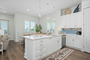 A kitchen with white cabinetry, a central island, and a dining area with a white table and chairs. - Coastal Charm | Pet Friendly | Steps to Beach (Port Aransas)