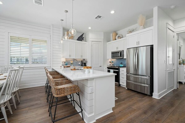 A modern kitchen with white cabinetry, a central island, bar stools, a stainless steel refrigerator, and a microwave. - Coastal Charm | Pet Friendly | Steps to Beach (Port Aransas)
