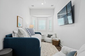 A modern living room with a blue sofa, a white armchair, and a small table. There is a framed picture on the wall and a window with blinds. - Coastal Charm | Pet Friendly | Steps to Beach (Port Aransas)