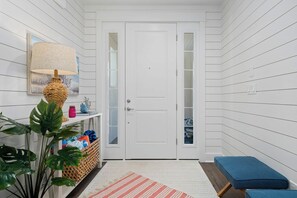 A hallway with white paneled walls, a white door, a wicker lamp, a plant, a bench, and a striped rug. - Coastal Charm | Pet Friendly | Steps to Beach (Port Aransas)