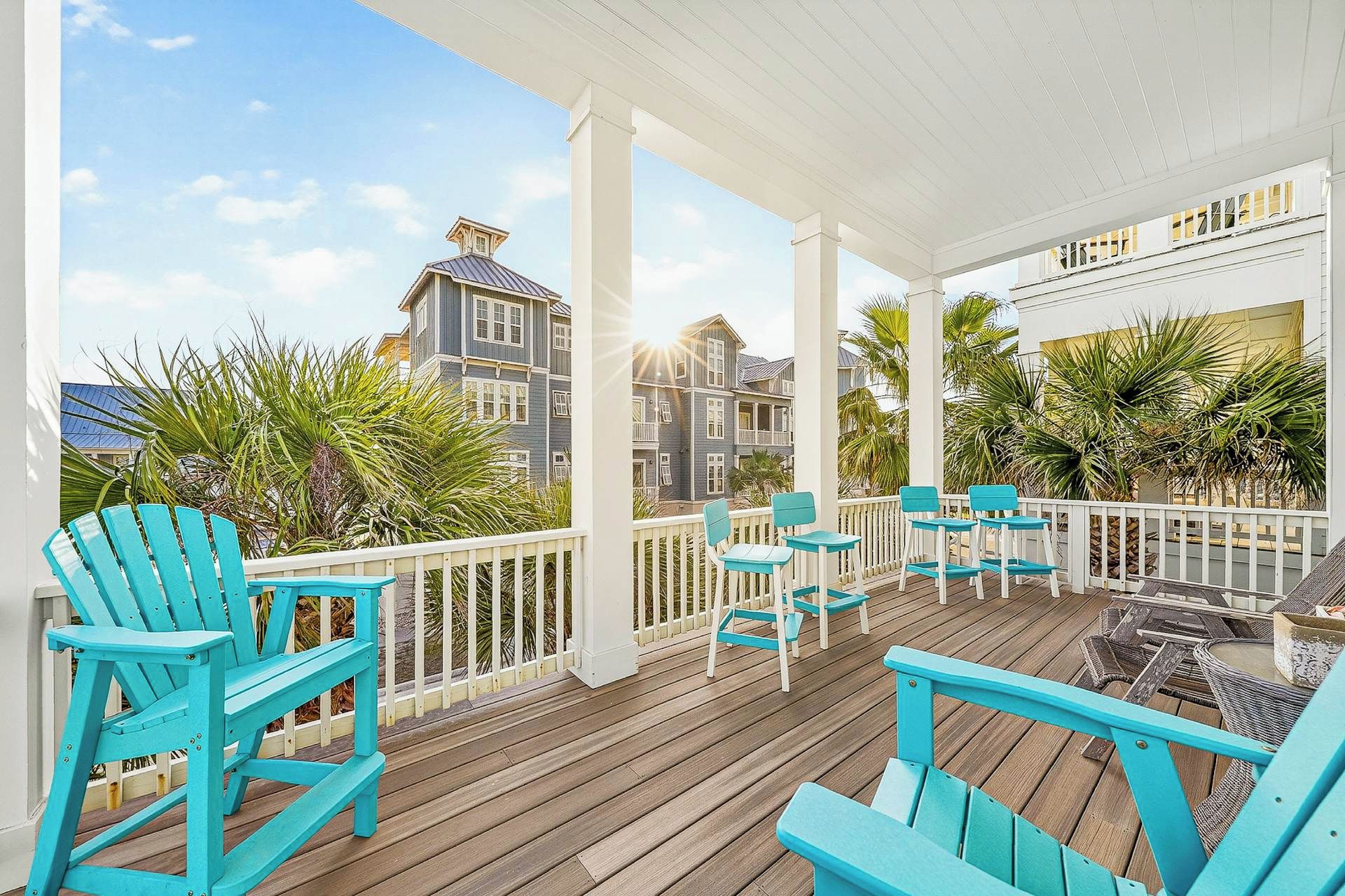 Una terraza de madera con sillas Adirondack turquesas y una mesita, con vistas a una zona residencial con un edificio blanco y palmeras.